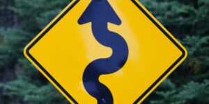 Yellow road sign with a winding arrow indicating a curvy road ahead, set against a background of green trees.