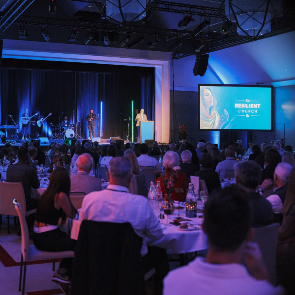 Audience seated at round tables watching a speaker on stage during a conference.