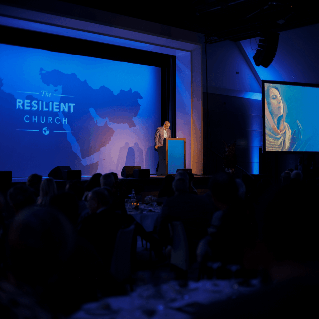 Speaker presenting on stage at a conference with a large screen and blue lighting.
